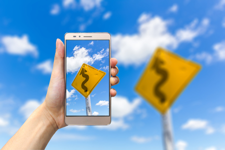 hand holding smartphone taking picture winding road sign with blue sky backgroundの写真素材