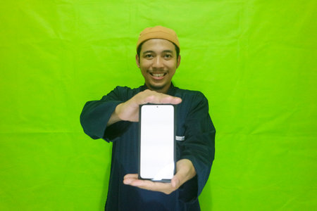Asian muslim young man showing smartphone with blank screen isolated on green backgroundの写真素材