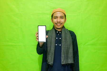 Asian muslim young man showing smartphone with blank screen isolated on green backgroundの写真素材