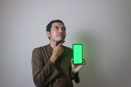 Happy Young Asian Muslim man showing blank green phone screen and pointing phone screen