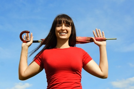 Young brunette girl in red shirt holding closed red umbrella behind head, blue skyの写真素材