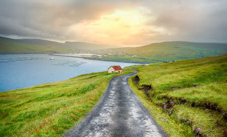 Beautiful trekking path at sunset with green meadows and views of the bay and Village of Sandavagur, Faroe Islands, Denmark, in the North Atlantic.の写真素材