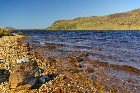 Lakeshore with pebbles, rocks in the foreground and hills in the distanceの写真素材