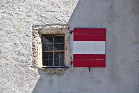 Rough plastered castle wall with window and wooden shutters colored like the Austrian flag, partly shaded and partly lit by sunlightの写真素材