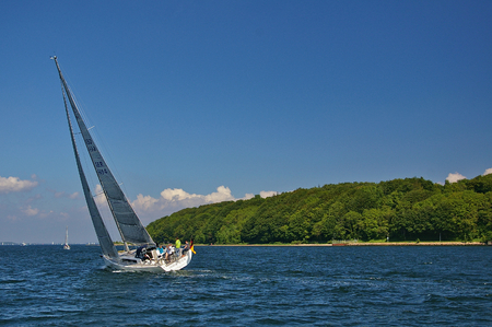 Flensburg, Germany - May 31st, 2011 - Modern sailing yacht in calm waters heeling over with blue sky and green wooded shoreline near Flensburg on the south shore of the Flensburg fiord.のeditorial素材