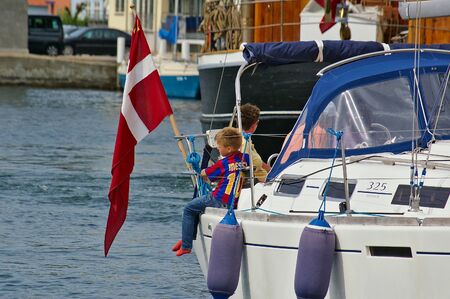 Sonderborg, Denmark - July 5th, 2012 - Young boy with soccer shirt sitting on the gunwhale of a white sailing yacht with the Danish flag next to him looking out over the waterのeditorial素材