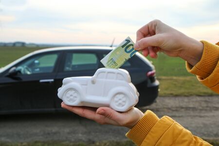 The hand is puting a banknote into the ceramic car shaped money box. The real car is behind. It can be symbol for costs for carÂ´s repairs, investment,の写真素材