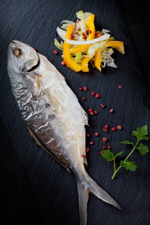 Baked trevally fish on stone plate, garnished with mixed vegetables salad, red pepper in grains and parsley, overhead shot.の写真素材