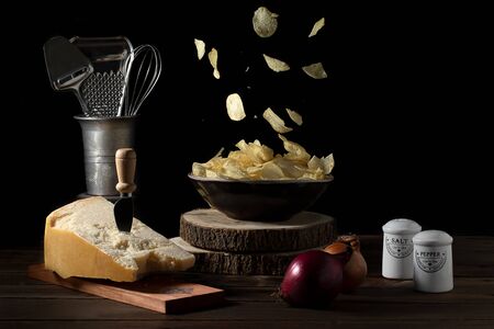 Potato chips falling in a bowl on a table with parmesan cheese, onions, salt and pepper - Dark mood