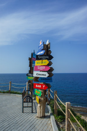 A vibrant signpost stands prominently near the shore, pointing towards various global destinations, with the serene sea creating a tranquil backdrop on a sunny day.の写真素材