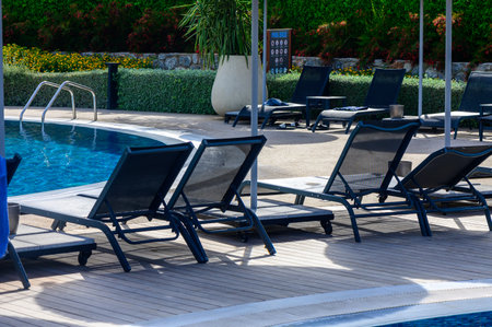 Lounge chairs are arranged beside a calm swimming pool on a warm sunny day, offering a perfect spot for relaxation among vibrant plants and trees.の写真素材