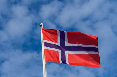 A vibrant Norwegian flag is seen fluttering against a backdrop of bright blue skies, representing...
