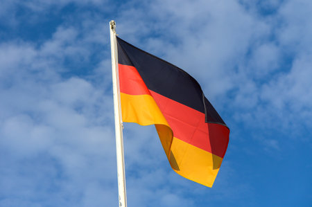 The German flag, with its distinctive black, red, and yellow stripes, flutters in the breeze under a clear blue sky filled with small clouds.