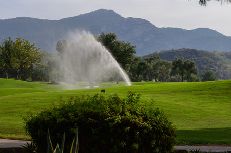 Morning light illuminates the tranquil golf course, where a fountain dances gracefully. Towering mountains and vibrant trees create a peaceful atmosphere.の写真素材