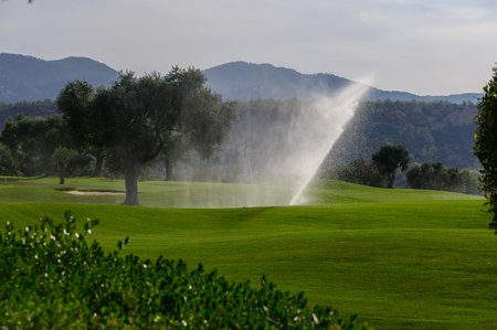 Beneath a clear sky, the serene golf course boasts vibrant green fairways, with sprinklers softly misting the landscape, enhancing the natural beauty of the surroundings.の写真素材