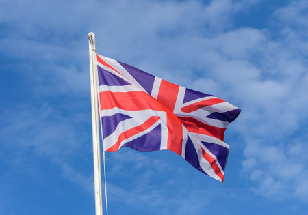 The Union Jack flutters in the gentle breeze, showing its bold colors against the backdrop of a bright, cloud-speckled sky. A true emblem of British identity.の写真素材
