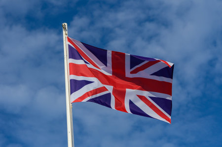 A vibrant Union Jack flag flutters atop a flagpole, set against a bright blue sky dotted with wispy...