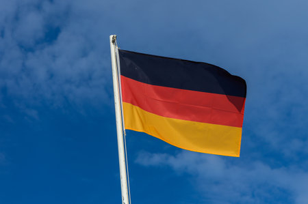 The German flag, featuring black, red, and gold stripes, flutters in the breeze against a bright...
