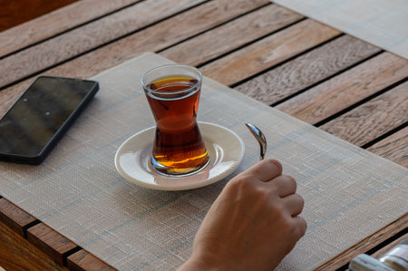 A hand delicately holds a teaspoon beside a small glass of tea resting on a plate, adding warmth to...