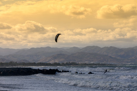 A kitesurfer skillfully maneuvers across the ocean waves as the sun sets behind mountain silhouettes.の写真素材