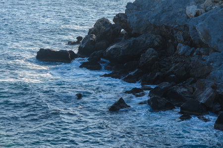 Gently rolling waves meet dark, jagged rocks as twilight casts a soft glow over the coastal...