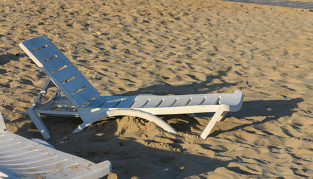 A damaged white lounge chair sits abandoned on the beach, casting shadows as the sun sets over the...