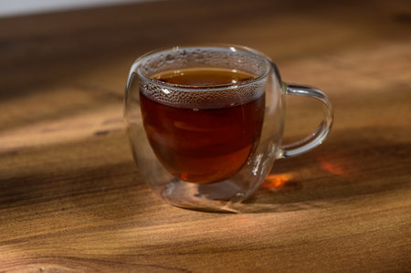Warmth radiates from a glass cup filled with rich tea, set against the inviting backdrop of wooden...