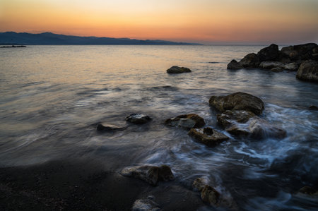 Golden hues blend into the horizon as gentle waves caress the rocky coast at dusk.の写真素材