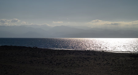 Gentle waves lap at the shore as the sun sets behind the distant mountains, casting a serene glow on...
