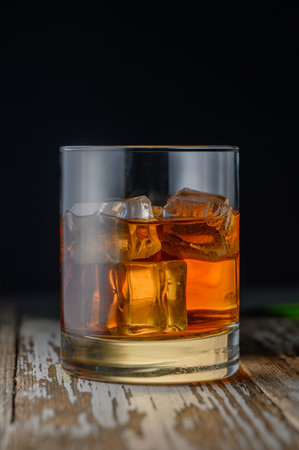 A glass filled with a golden drink and ice sits on a rustic wooden table, glistening under soft light.の写真素材