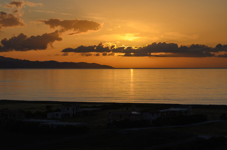 Golden sunlight reflects on calm waters as the sun descends behind distant hills, creating a peaceful twilight atmosphere along the tranquil coastline. The beauty of nature inspires serenity.の写真素材