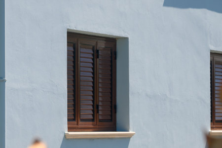 Bright sunlight highlights well-crafted wooden shutters on a pastel blue wall of a coastal home. The scene exudes tranquility and captures the essence of relaxed living by the sea.の写真素材