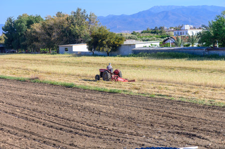 A farmer plowing a field with a tractor near a village and mountains in Northern Cyprus. Outdoor rural landscape showcasing traditional Mediterranean agriculture.の写真素材