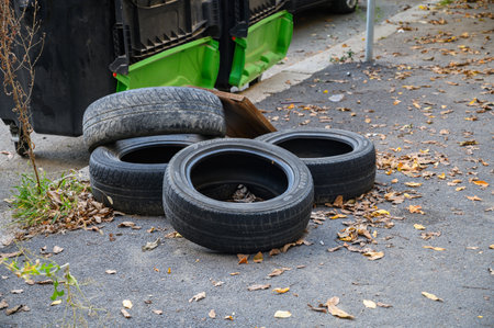 Several old car tires lie near garbage containers on a city street. The scene represents environmental pollution, illegal waste disposal, and the global problem of tire recycling and waste management.の写真素材
