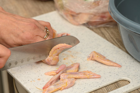 Close-up of a woman in a white apron carefully slicing fresh chicken wings on a wooden board on the kitchen table. Salt, pepper, juice droplets, and warm morning light create a lively backgroundの写真素材