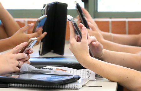 Students holding and using a range of digital devices. Mobile cell phones tablets and technology being handled by students at a desk in a school educational settingのeditorial素材