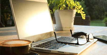Beautiful golden morning sunshine on an outdoor table setting with hot coffee, a laptop computer, mobile cell phone, glasses and mouse ready for online shopping hecking emails working from home.の写真素材