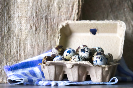 A large carton of several quail eggs, on a cloth and wooden board background.