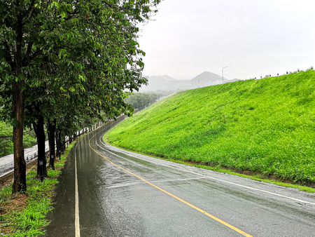 Near the embankment of a large reservoir, a soaking wet, tree lined, Thai countryside road goes downwards into the distance landscape, during a tropical downpour.の写真素材