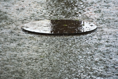 An interesting sight of a round, flooded, manhole cover, set during a hard tropical thunderstorm in Thailand, where raindrops and debris cover this sewer lid, in a somewhat beautiful way.の写真素材