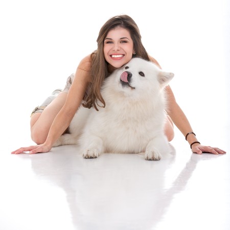 a studio image of a young woman and her white dog, with her sitting on her hands and knees on top of the dog, looking forward, smiling and the dog looking on a side, licking it's mouth.の写真素材