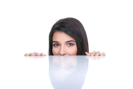 a portrait of a young woman, posing on a white background. she only has the tips of her fingers on a white table and only her eyes and nose are showing from behind the table. she has a smile on her face and a childish expression.の写真素材