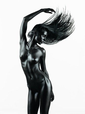 artistic nude of a young woman with black painted skin on white background, in a dance movement with her hand up in the airの写真素材