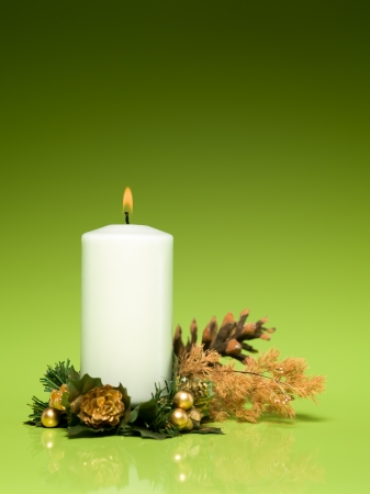 closeup of single white burning candle with cone, bracnhes and artificial leaves arround it on green gradient backgroundの写真素材
