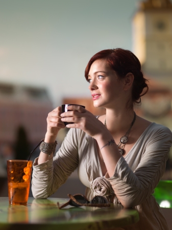 young gorgeaous caucasian red haired girl sitting outside in the city at a table of a cafe drinking coffe and looking at somethingの写真素材