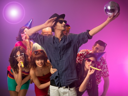 close-up of young handsome caucasian man with sunglasses holding a disco ball and looking at it, with other party people behind him surounded by colorful lightsの写真素材