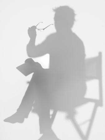 man sitting on director's chair reading a book with eyeglasses in his hand, behind a diffuse surfaceの写真素材