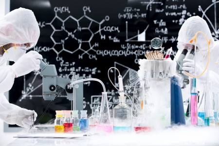 side-view of two people manipulating laboratory tools and colorful liquids on a lab table with a blackboard on the backgroundの写真素材