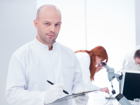 close-up of a smiling scientist holding a clip-board in a chemistry lab around a worktable where two...