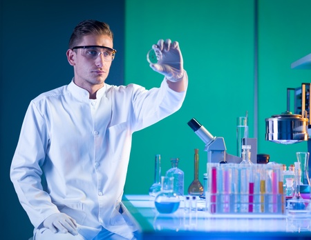 close-up of scientist sitting down near a laboratory table with chemistry glassware on top, holding...